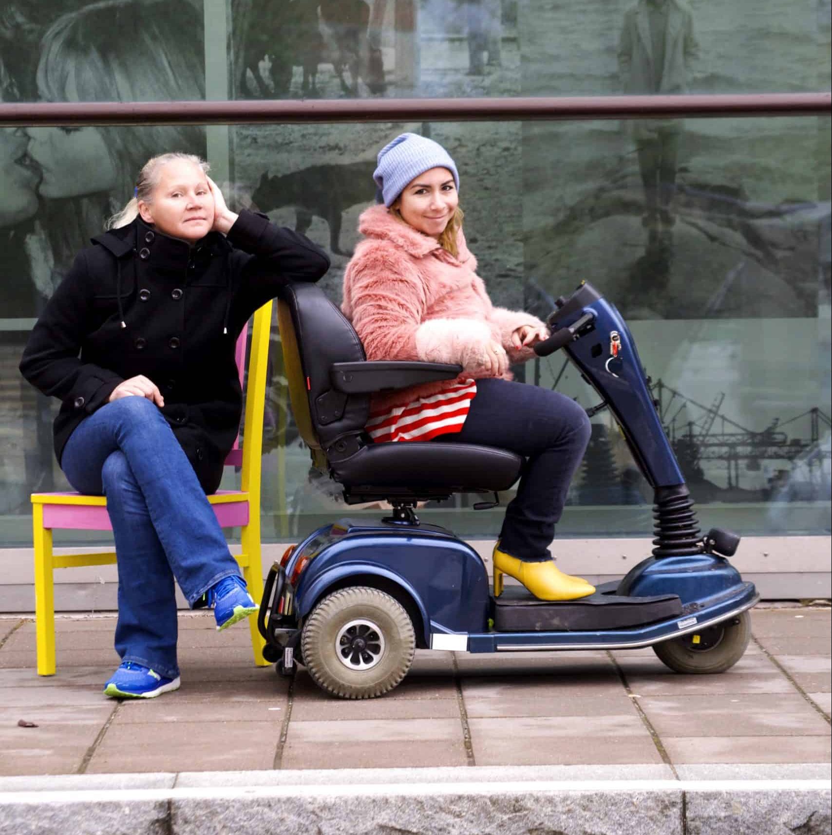 Person i rullstol och kvinna sittande på stol utomhus.