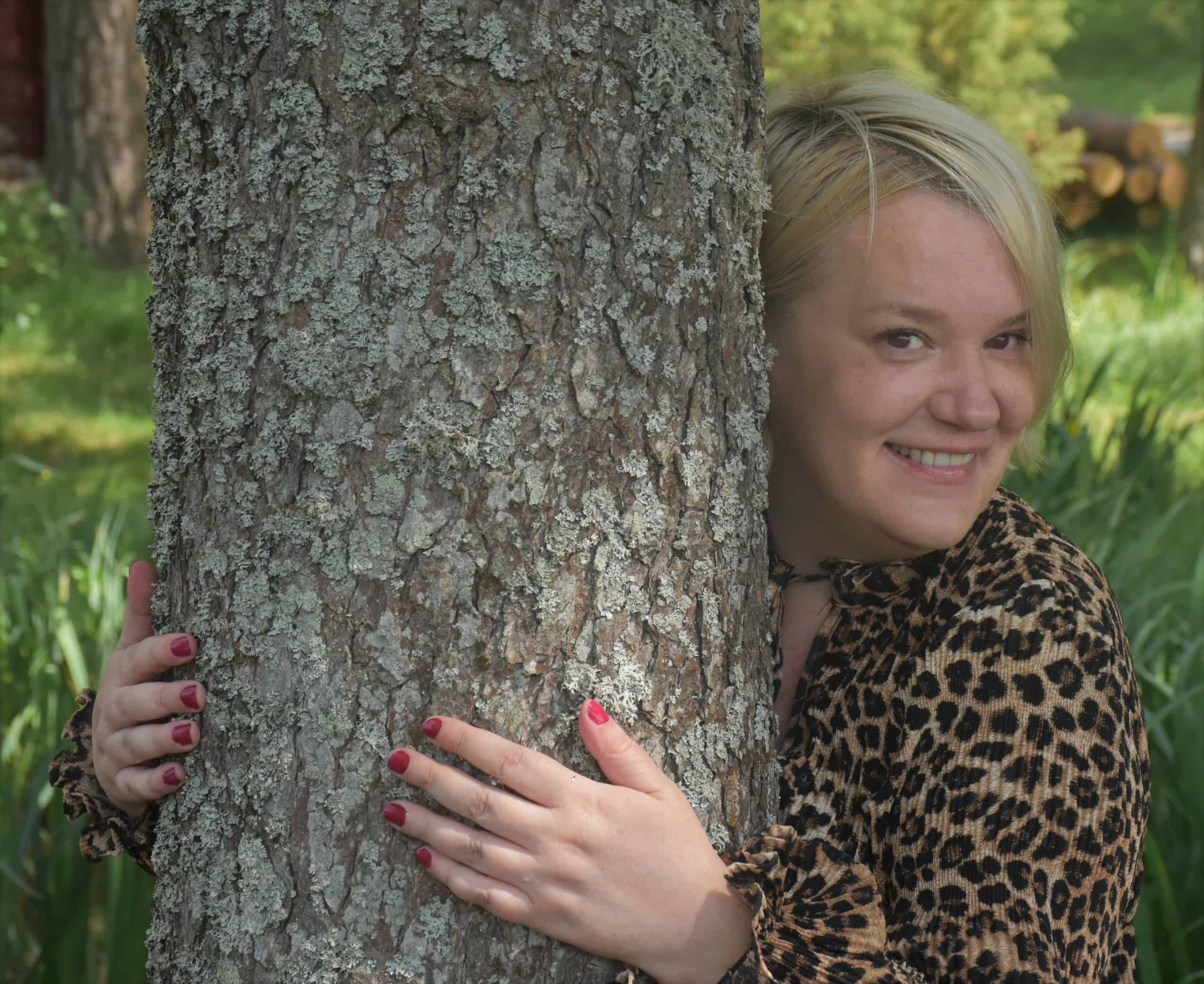 En kvinna omfamnar ett träd i en grön parkmiljö, ler och ser glad ut.