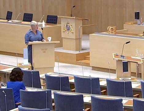 Politiker håller tal i ett mötesrum med tomma stolar och mikrofoner.