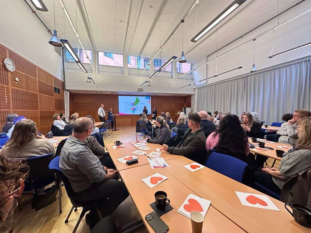 Konferens i stort mötesrum med presentation och deltagare.