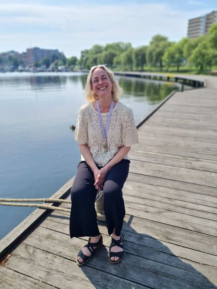 Venus sittande på en träbrygga vid vattnet i en park, med gröna träd och stad i bakgrunden.