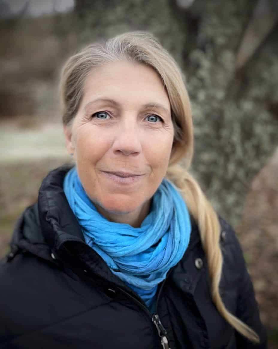 En kvinna med blont hår och blå scarf utomhus, ler vänligt.